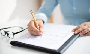 Dłoń pisze złotym długopisem na dokumencie, który wygląda jak aneks do umowy o pracę. Obok leżą okulary.