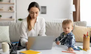 Kobieta pracuje na laptopie, podczas gdy jej syn rysuje obok. To codzienna opieka na dziecko i praca zdalna.