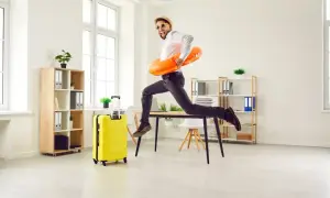 Radosny mężczyzna w okularach i kapeluszu skacze z biurka z kołem ratunkowym, obok żółta walizka. Czas na urlop zgodny z kodeksem pracy!