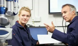 Młoda pracownica słucha wskazówek starszego kolegi podczas omawiania dokumentacji technicznej. To część regularnych badań okresowych pracowników.