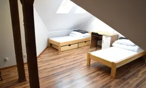 Pokój w bursie z dwoma łóżkami, biurkiem i szafką. Drewniane belki i podłoga nadają przytulny klimat.