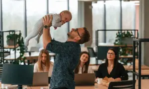 Szczęśliwy ojciec podrzuca dziecko w biurze, podczas gdy koleżanki pracują. Czy urlop wychowawczy wlicza się do stażu pracy?