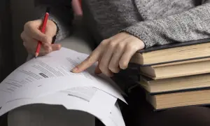 Ręka z ołówkiem wskazuje na dokumenty, stos książek obok. Czekanie na wyniki matur bywa stresujące.