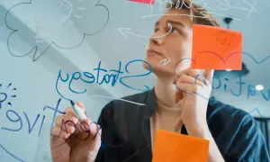 Młody mężczyzna pisze na szkle, tworząc schemat. Widać żarówkę symbolizującą pomysł i słowo "hit".
