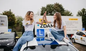 Dwie uśmiechnięte dziewczyny w koszulkach "Ja Zdam Szkoła Jazdy" świętują sukces, siedząc na dachu samochodu. To znak, że zdały egzamin klasyfikacyjny!