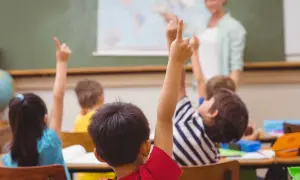 Uczniowie z podniesionymi rękami, gotowi do odpowiedzi. Nauczycielka ocenia ich zaangażowanie, co wpłynie na oceny z zachowania.