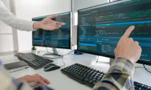 Dwóch programistów analizuje kod na monitorach. Jeden wskazuje palcem na ekran, drugi gestykuluje.