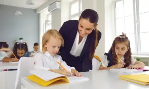 Nauczycielka podczas hospitacji wspiera ucznia w pisaniu. Dzieci siedzą przy ławkach, skupione na zadaniu.
