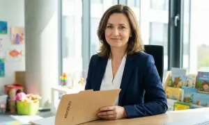Kobieta w granatowej marynarce trzyma teczkę z napisem "PROCEDURY". W tle przedszkolne rysunki i książki. To przykładowy opis oceny pracy nauczyciela przedszkola.