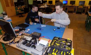 Dwóch uczniów ćwiczy technikę informatyczną, montując kable sieciowe. Na stole sprzęt i narzędzia.