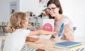 Uśmiechnięta psycholog z okularami pokazuje dziecku karty z buźkami. To część przygotowania pedagogicznego dla psychologów.