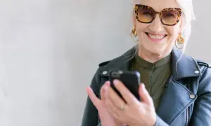 Uśmiechnięta starsza pani w okularach w panterkę sprawdza telefon. Może szuka informacji o kodeksie pracy i odprawie emerytalnej.