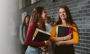 Dwie uczennice rozmawiają, trzymając książki. Może omawiają przeniesienie nauczyciela do innej szkoły i nowe wyzwania.