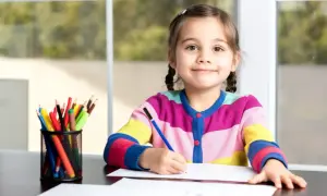 Uśmiechnięta dziewczynka z warkoczami rozwija swoją percepcję wzrokową, rysując kredkami przy oknie.