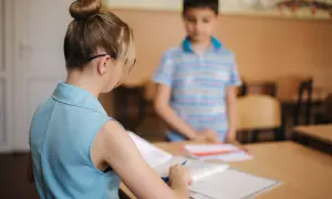 Nauczycielka z kucykiem przygląda się zeszytom, podczas gdy uczeń stoi w tle. Czy to zapowiedź podwyżki dla nauczycieli? Najnowsze wiadomości wkrótce.
