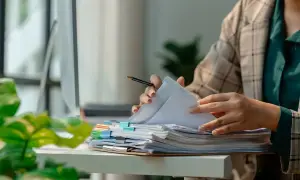 Pracownik przegląda dokumenty, marząc o urlopie dla poratowania zdrowia. Stos papierów czeka na biurku.