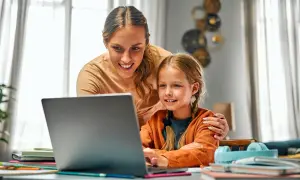 Uśmiechnięta mama pomaga córce w nauce przy laptopie. To przykład troskliwej opieki na dziecko 2 dni w tygodniu, gdy rodzice pracują.