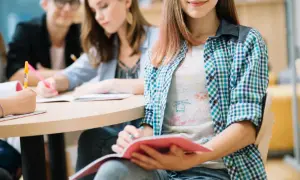 Uczniowie przy stole, skupieni na nauce biologii. Jedna z dziewczyn notuje, druga czyta, trzecia uśmiecha się, trzymając zeszyt.
