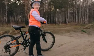 Dziewczynka w kasku i kamizelce przygotowuje się do egzaminu na kartę rowerową, stojąc obok swojego roweru na leśnej ścieżce.