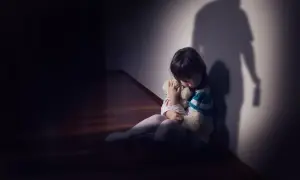 Dziecko tuli misia, skulone w kącie. Cień dorosłego z butelką sugeruje agresję i strach.