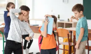 Chłopiec zasłania się zeszytem, podczas gdy inni uczniowie w klasie wydają się być zaangażowani w bullying.