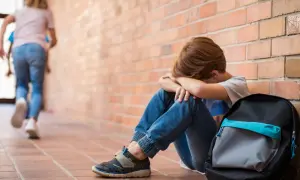 Chłopiec siedzi przy ceglanej ścianie, przytulając się do siebie. W tle inni uczniowie biegną, ignorując jego cierpienie. To obraz mobbingu w szkole.