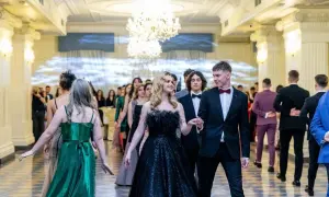 Młoda para idzie na studniówkę. Dziewczyna w czarnej, błyszczącej sukni z piórami, chłopak w eleganckim garniturze. W tle inni uczniowie w balowych strojach.