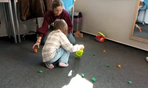 Dzieci bawią się na dywanie, zbierając małe, kolorowe figurki. W tle widać dorosłą osobę. To świetna zabawa z elementami wwr.