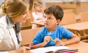 Nauczycielka wspiera chłopca z **specjalnymi potrzebami edukacyjnymi** w nauce. W tle widać inne dzieci.