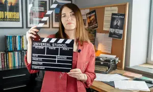 Kobieta trzyma klaps filmowy, gotowa do rozpoczęcia zajęć. W tle plakaty filmowe i maszyna do pisania, tworząc atmosferę pracy nad scenariuszem.