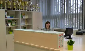 Kobieta siedzi za ladą w sekretariacie, wśród licznych pucharów i dyplomów.