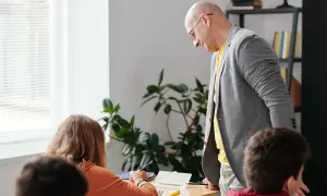 Nauczyciel pochyla się nad uczennicą, która pisze. W tle roślina i regał z książkami. Czy to początek choroby zawodowej nauczycieli?