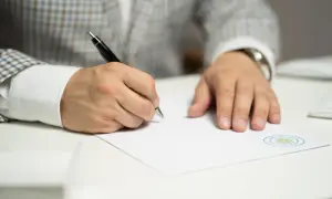Mężczyzna w eleganckim garniturze dokonuje sprostowania świadectwa pracy, pisząc długopisem na białej kartce z firmowym logo.