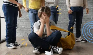 Chłopiec siedzi na podłodze, zakrywając twarz dłońmi, podczas gdy grupa dzieci stoi nad nim. To obraz tego, czym jest **bullying**.