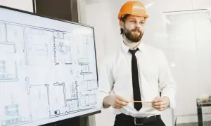 Mężczyzna w kasku pomarańczowym prezentuje projekt domu na ekranie. To szkolenie BHP dla pracownika, który zapoznaje się z planem.