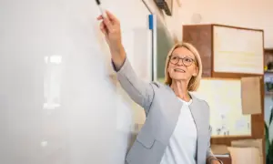 Uśmiechnięta nauczycielka z markerem w dłoni, gotowa do oceny pracy uczniów przy tablicy.