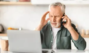 Zmartwiony mężczyzna rozmawia przez telefon, trzymając się za głowę. Obok laptopa i kubka. Wygląda na to, że jego urlop z powodu siły wyższej został zakłócony.