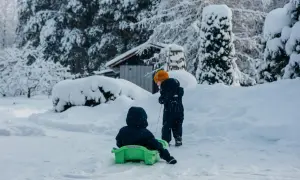Dzieci cieszą się śnieżnymi feriami małopolskimi. Jedno zjeżdża na sankach, drugie ciągnie je przez zasypany śniegiem ogród.