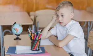 Chłopiec w białej koszuli siedzi przy biurku z globusem i kredkami. Zamyślony, może czeka na odroczenie obowiązku szkolnego.