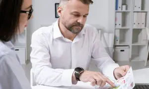 Mężczyzna z zarostem i zegarkiem na ręku analizuje wykresy, podczas gdy kobieta w okularach obserwuje. Ich praca wymaga ścisłego nadzoru.