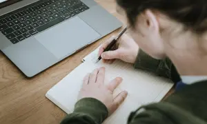 Nauczyciel pisze opinię o uczniu w zeszycie, obok leży laptop.