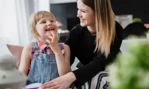 Uśmiechnięta dziewczynka z niepełnosprawnością sprzężoną bawi się z mamą, która wspiera ją w nauce.
