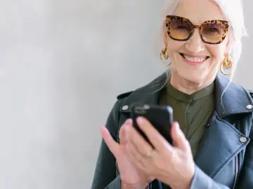 Uśmiechnięta starsza pani w okularach w panterkę sprawdza telefon. Może szuka informacji o kodeksie pracy i odprawie emerytalnej.