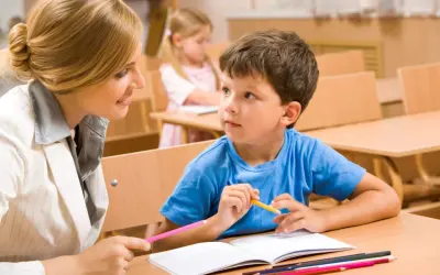 Nauczycielka pomaga chłopcu w odrabianiu pracy domowej. Dziecko uważnie słucha, trzymając kredkę.