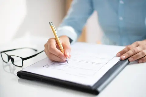 Dłoń pisze złotym długopisem na dokumencie, który wygląda jak aneks do umowy o pracę. Obok leżą okulary.