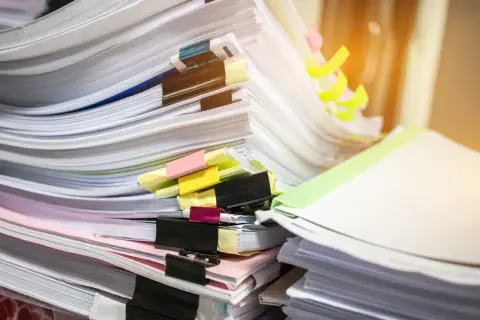 Stosy dokumentów, wśród nich duplikat świadectwa pracy, z kolorowymi zakładkami i spinaczami.