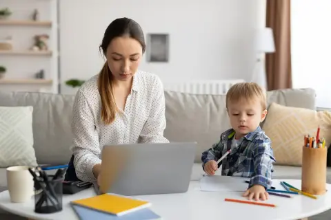 Kobieta pracuje na laptopie, podczas gdy jej syn rysuje obok. To codzienna opieka na dziecko i praca zdalna.