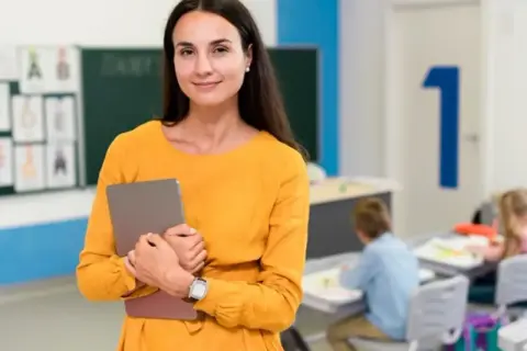Nauczycielka z tabletem, uśmiechnięta, w klasie z dziećmi. Posiada kwalifikacje nauczyciela wspomagającego.