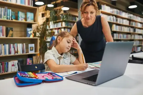 Dziewczynka z ołówkiem w dłoni, zamyślona nad zadaniem, przygotowuje podanie do szkoły średniej. Nauczycielka wspiera ją w bibliotece.