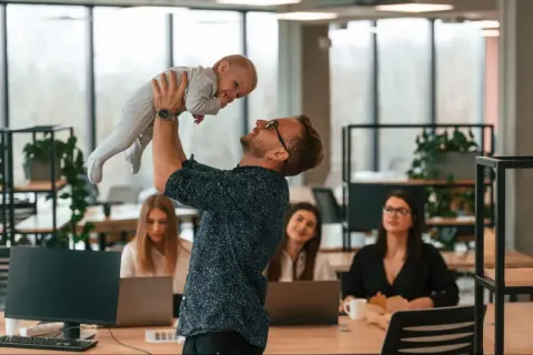 Szczęśliwy ojciec podrzuca dziecko w biurze, podczas gdy koleżanki pracują. Czy urlop wychowawczy wlicza się do stażu pracy?